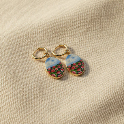 A pair of handmade gold hoop earrings with oval charms featuring a hand-painted watercolor style landscape of red flowers under a blue sky, displayed on a textured beige fabric background. The hoops are PVD 18k Gold Coated Titanium.