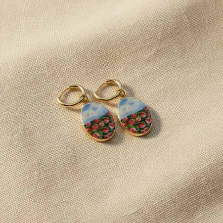 A pair of handmade gold hoop earrings with oval charms featuring a hand-painted watercolor style landscape of red flowers under a blue sky, displayed on a textured beige fabric background. The hoops are PVD 18k Gold Coated Titanium.