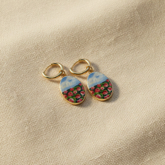 A pair of handmade gold hoop earrings with oval charms featuring a hand-painted watercolor style landscape of red flowers under a blue sky, displayed on a textured beige fabric background. The hoops are PVD 18k Gold Coated Titanium.
