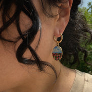 A close-up side view of a person wearing a gold hoop earring with an oval drop charm featuring a colorful landscape scene, set against an outdoor background with green foliage.