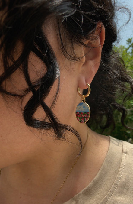 A close-up side view of a person wearing a gold hoop earring with an oval drop charm featuring a colorful landscape scene, set against an outdoor background with green foliage.