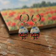A pair of handmade gold hoop earrings with oval drop charms featuring a split design: the top half shows a blue sky with clouds, and the bottom half shows a close-up field of red and pink flowers. The earrings are displayed on a rustic wooden surface with a blurred background showing a matching flower field and a book.