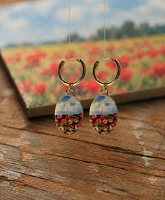 A pair of handmade gold hoop earrings with oval drop charms featuring a split design: the top half shows a blue sky with clouds, and the bottom half shows a close-up field of red and pink flowers. The earrings are displayed on a rustic wooden surface with a blurred background showing a matching flower field and a book.