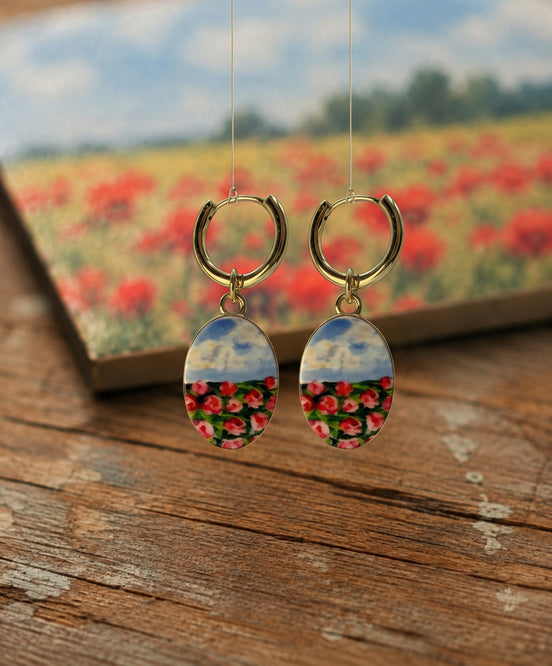 A pair of handmade gold hoop earrings with oval drop charms featuring a split design: the top half shows a blue sky with clouds, and the bottom half shows a close-up field of red and pink flowers. The earrings are displayed on a rustic wooden surface with a blurred background showing a matching flower field and a book.