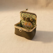 An open vintage-style kissel case with a textured olive green exterior, gold-tone metal frame, and a floral green-and-white patterned interior. Inside, a pair of round gold-tone earrings with floral enamel details rests on a beige fabric surface.