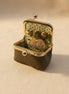An open vintage-style kissel case with a textured olive green exterior, gold-tone metal frame, and a floral green-and-white patterned interior. Inside, a pair of round gold-tone earrings with floral enamel details rests on a beige fabric surface.