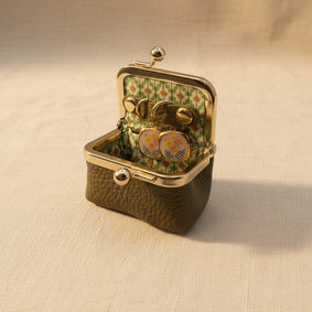 An open vintage-style kissel case with a textured olive green exterior, gold-tone metal frame, and a floral green-and-white patterned interior. Inside, a pair of round gold-tone earrings with floral enamel details rests on a beige fabric surface.