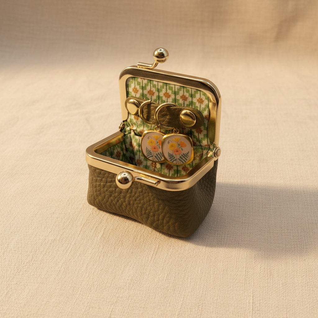 An open vintage-style kissel case with a textured olive green exterior, gold-tone metal frame, and a floral green-and-white patterned interior. Inside, a pair of round gold-tone earrings with floral enamel details rests on a beige fabric surface.