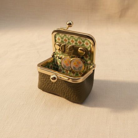 An open vintage-style kissel case with a textured olive green exterior, gold-tone metal frame, and a floral green-and-white patterned interior. Inside, a pair of round gold-tone earrings with floral enamel details rests on a beige fabric surface.