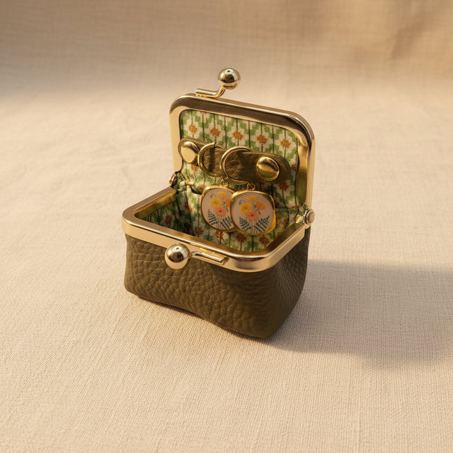 An open vintage-style kissel case with a textured olive green exterior, gold-tone metal frame, and a floral green-and-white patterned interior. Inside, a pair of round gold-tone earrings with floral enamel details rests on a beige fabric surface.